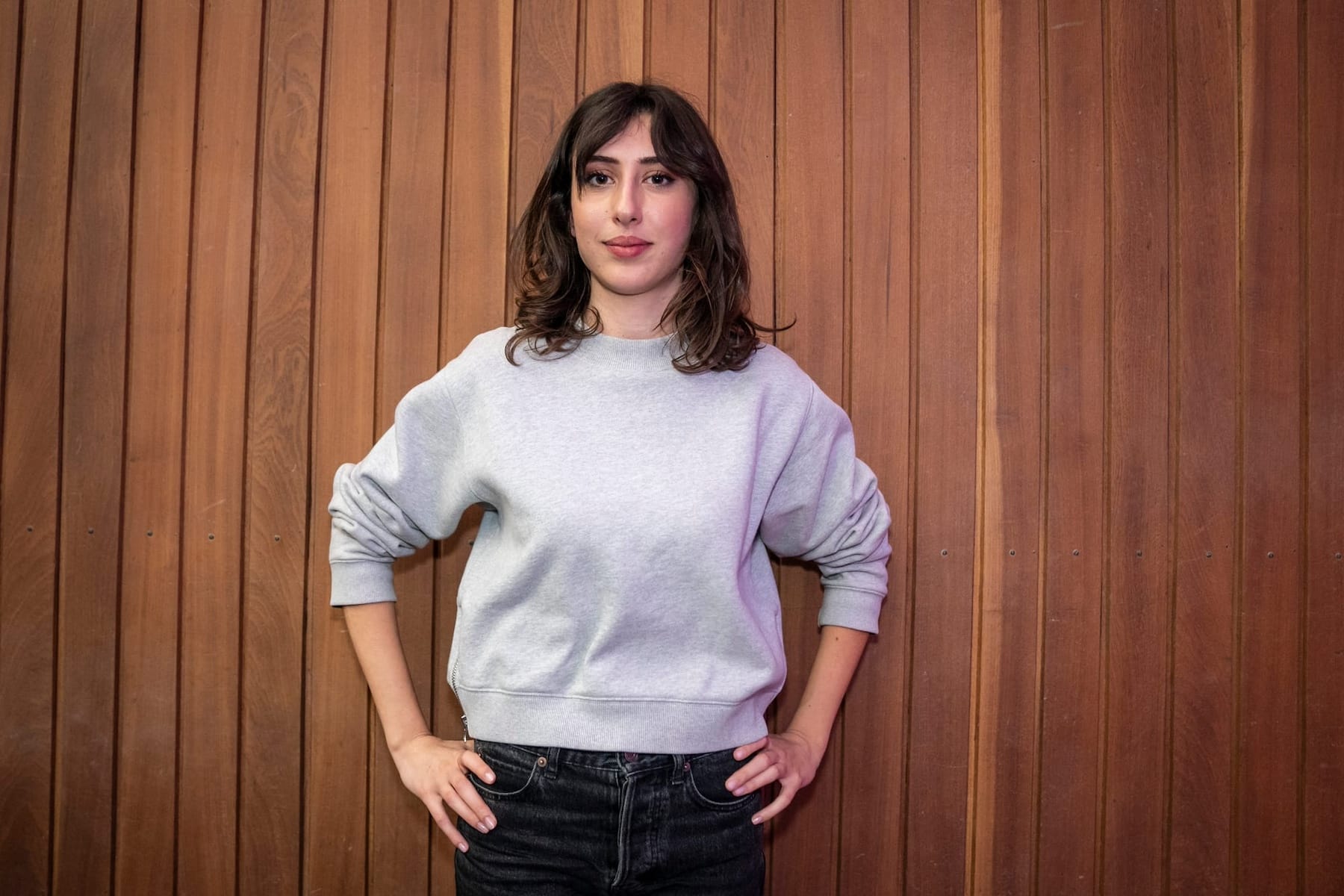 Sala is standing before a wood plank wall in daily outfit and looking into the camera with a confident smile.