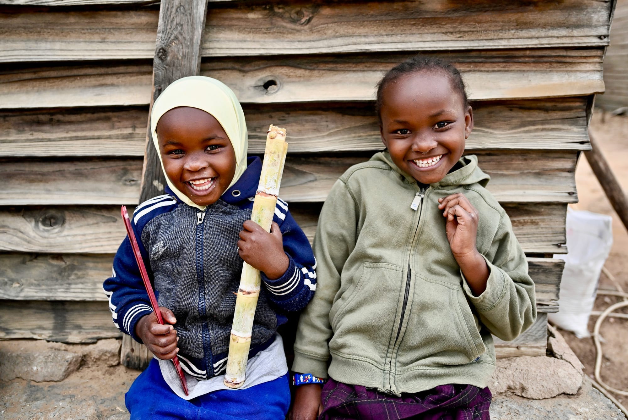 Two cheerful children smile at the camera in Mutoko, Zimbabwe, sitting before a wooden wall and holding sugarcane.