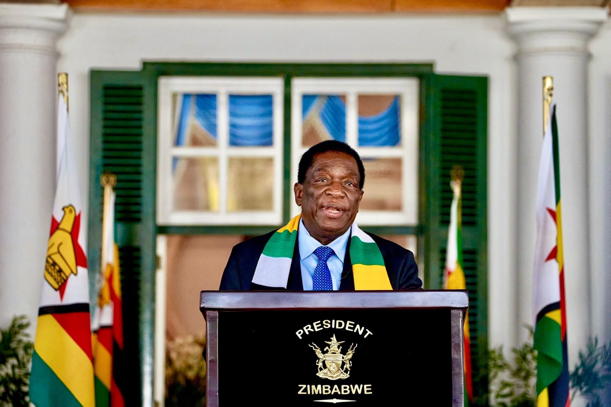 Zimbabwean President Emmerson Mnangagwa speaks at a press conference at State House, standing before a podium with the presidential emblem, flanked by Zimbabwean flags.