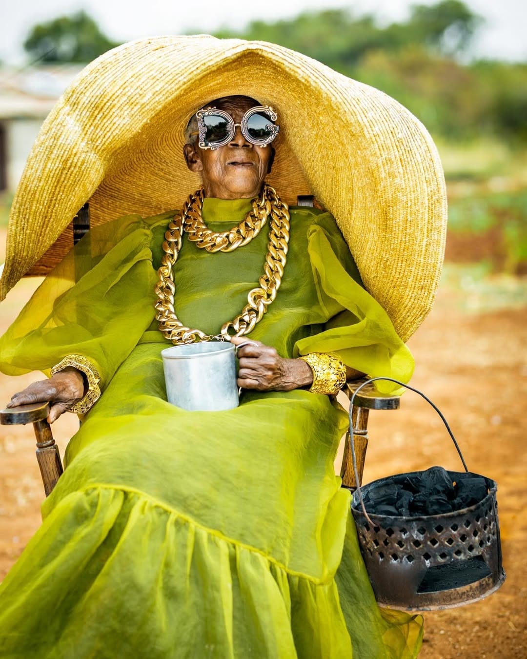 Chola is draped in a flowing lime-green gown with a massive straw hat. She wears bold, gold chain jewelry and oversized designer sunglasses while holding a big tin cup and a charcoal basket filled with coal.