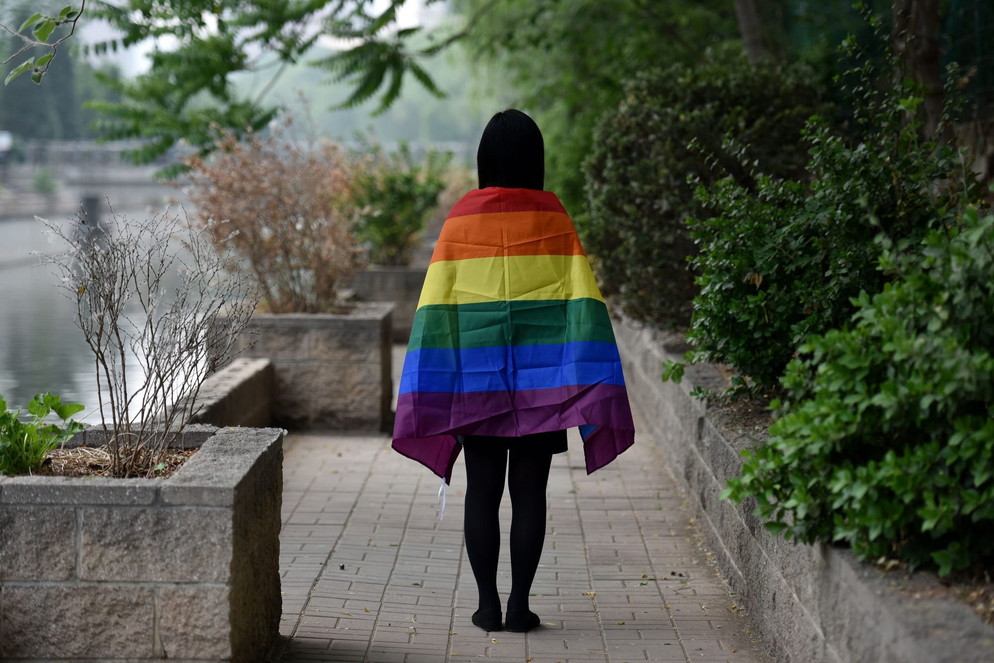 A person draped in a rainbow LGBTQ+ pride flag stands on a pathway in China, facing away from the camera, symbolizing the challenges and restrictions faced by the LGBTQ+ community in the country.