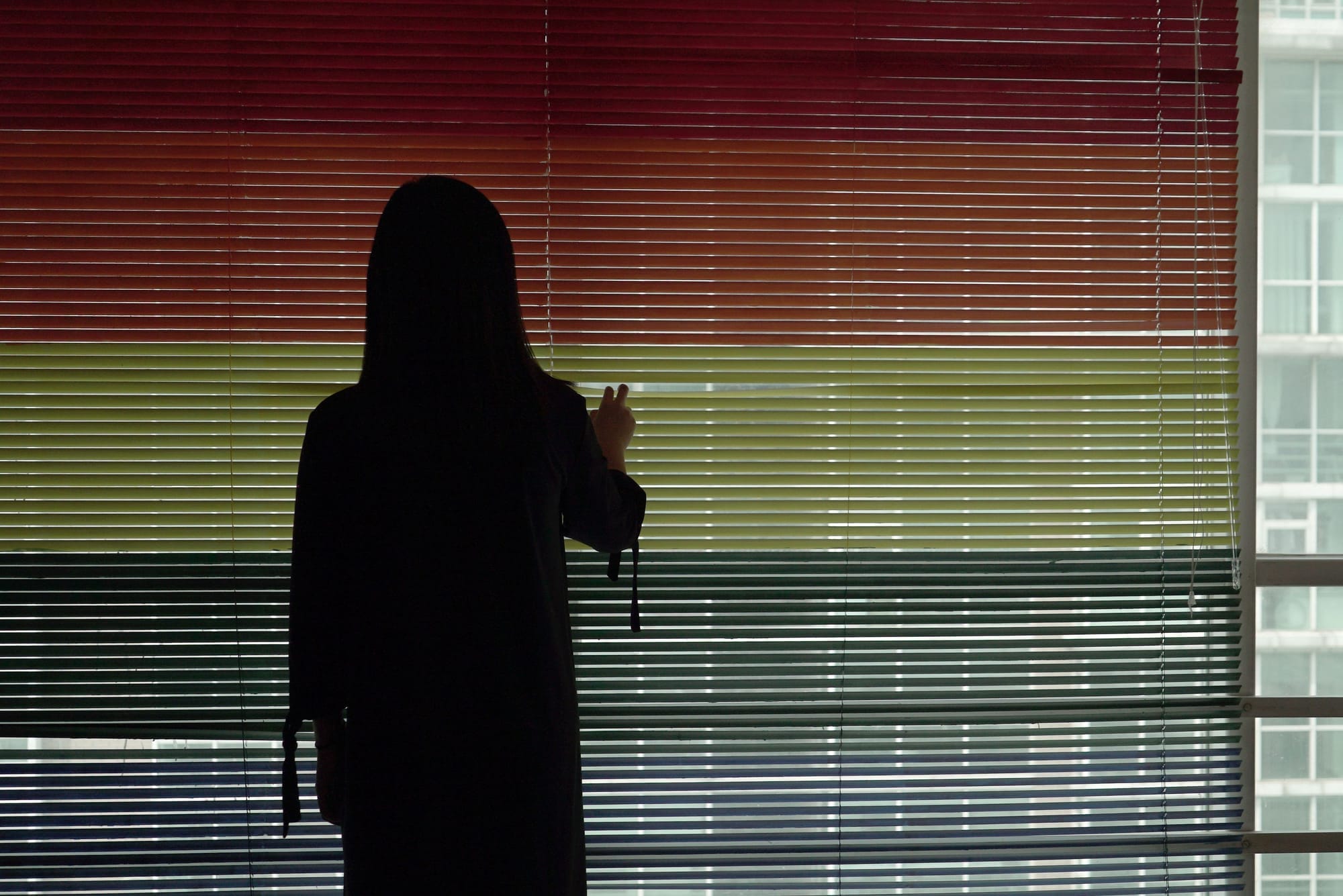 A gay student is standing behind rainbow blinds, turning away from the camera.