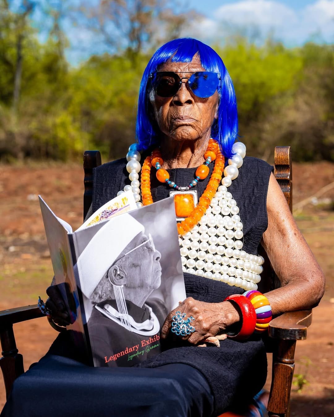 Chola is sitting in a wooden chair, wearing a blue wig, mirrored sunglasses, and colorful orange and white bead necklaces. She is flipping open a magazine featuring her own portrait on the cover, titled Legendary Exhibit and captioned Legendary Glamma.