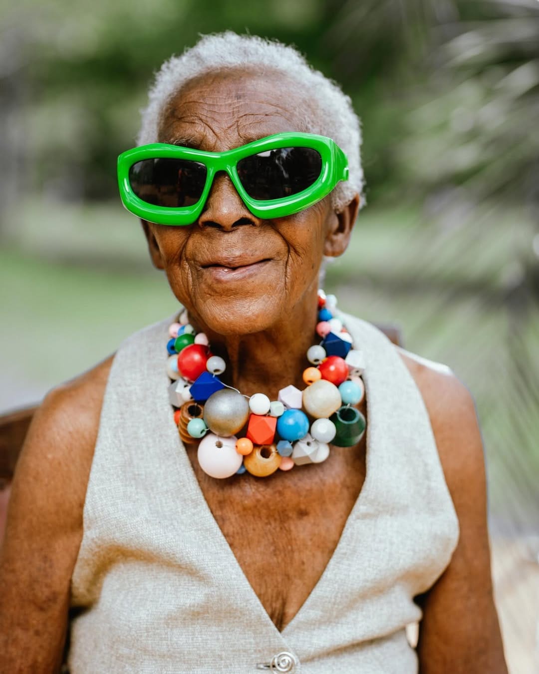 A close-up portrait of Chola wearing bold green oversized sunglasses, a white cotton waistcoat and a colorful chunky bead necklace. She smiles warmly with a confident expression.