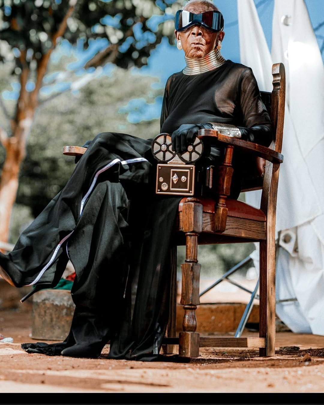 Chola sits on a wooden chair, wearing a pair of futuristic sunglasses, a black sheer outfit, and metallic accessories. She is also holding a vintage film reel camera.