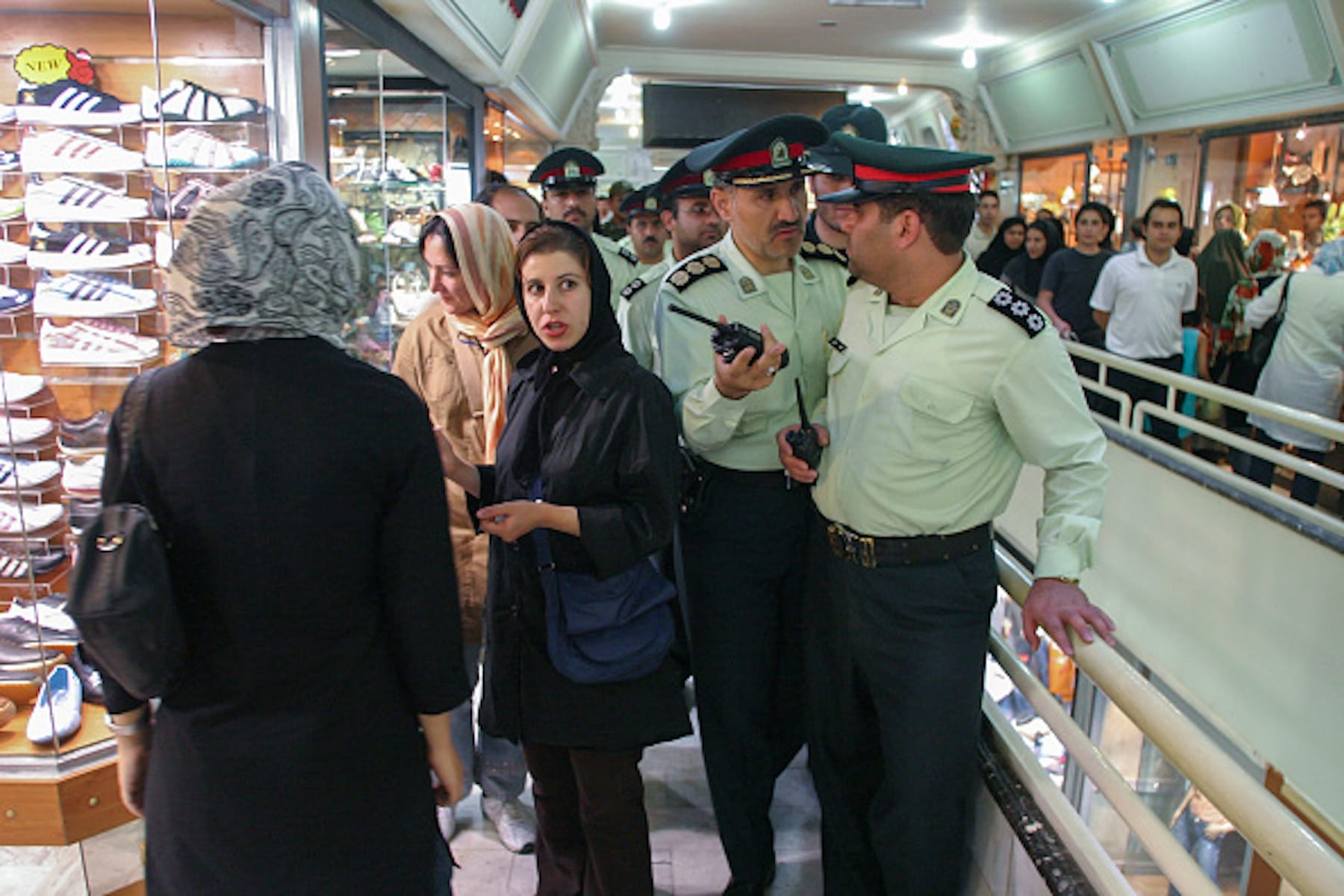 Iranian morality police officers in uniform monitor women in a shopping center, engaging with them while standing near a shoe store, amidst other shoppers in the background.