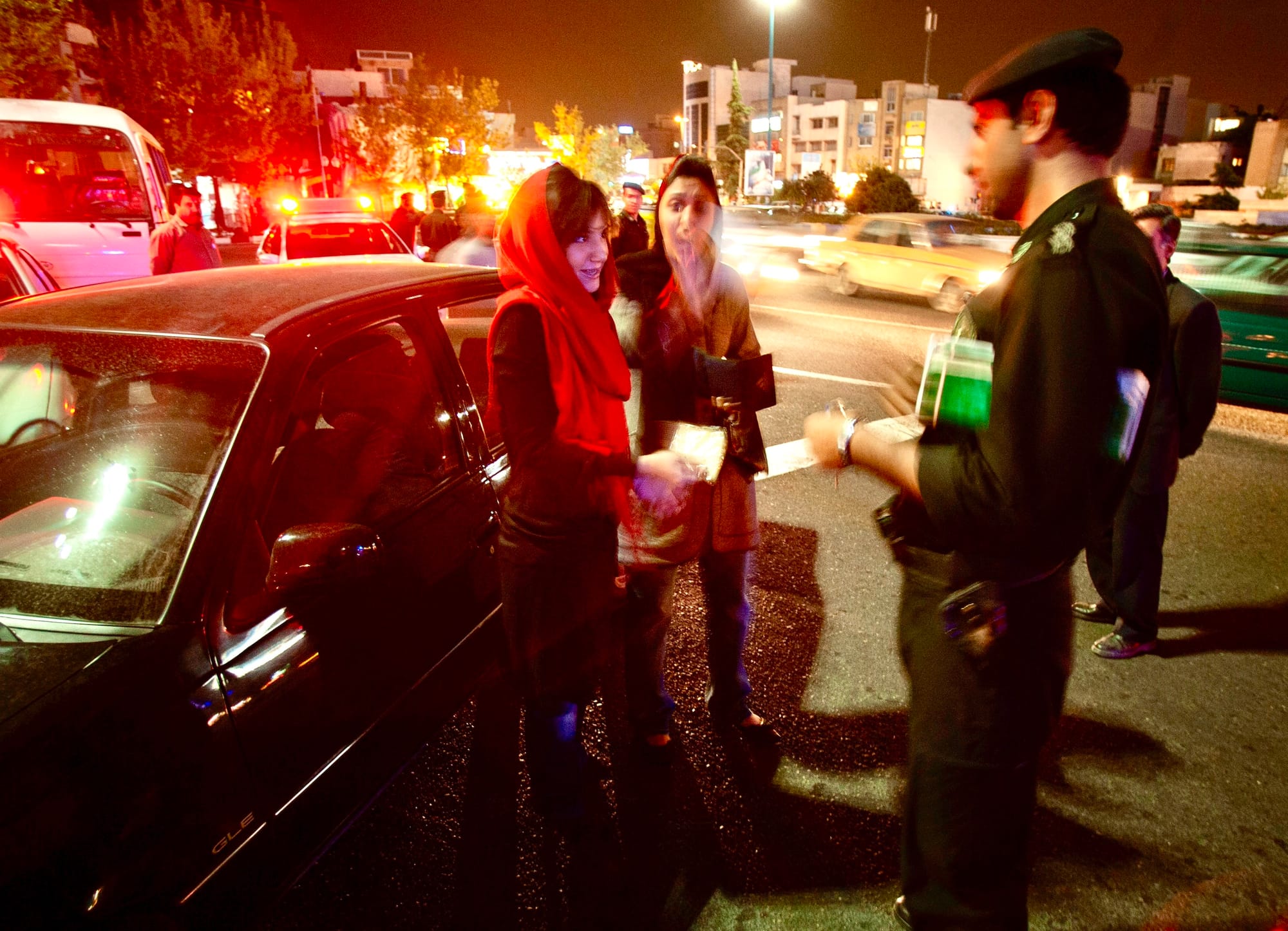 Iranian morality policeman speaks to two women, one wearing a red scarf covering her hair, beside a car at night in the middle of traffic.