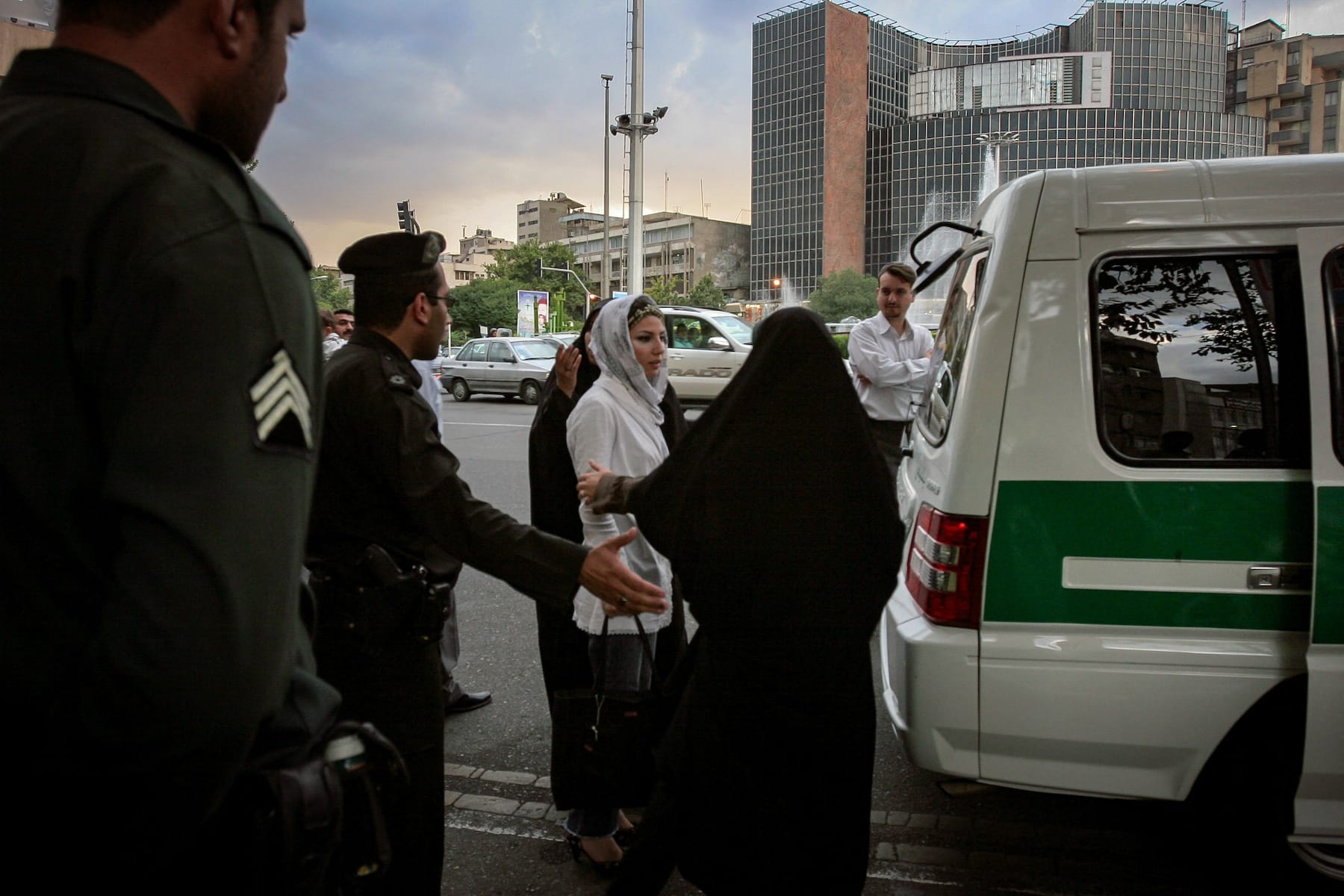 Iranian morality police officers detain a woman in a white headscarf by a police vehicle in a busy urban area.