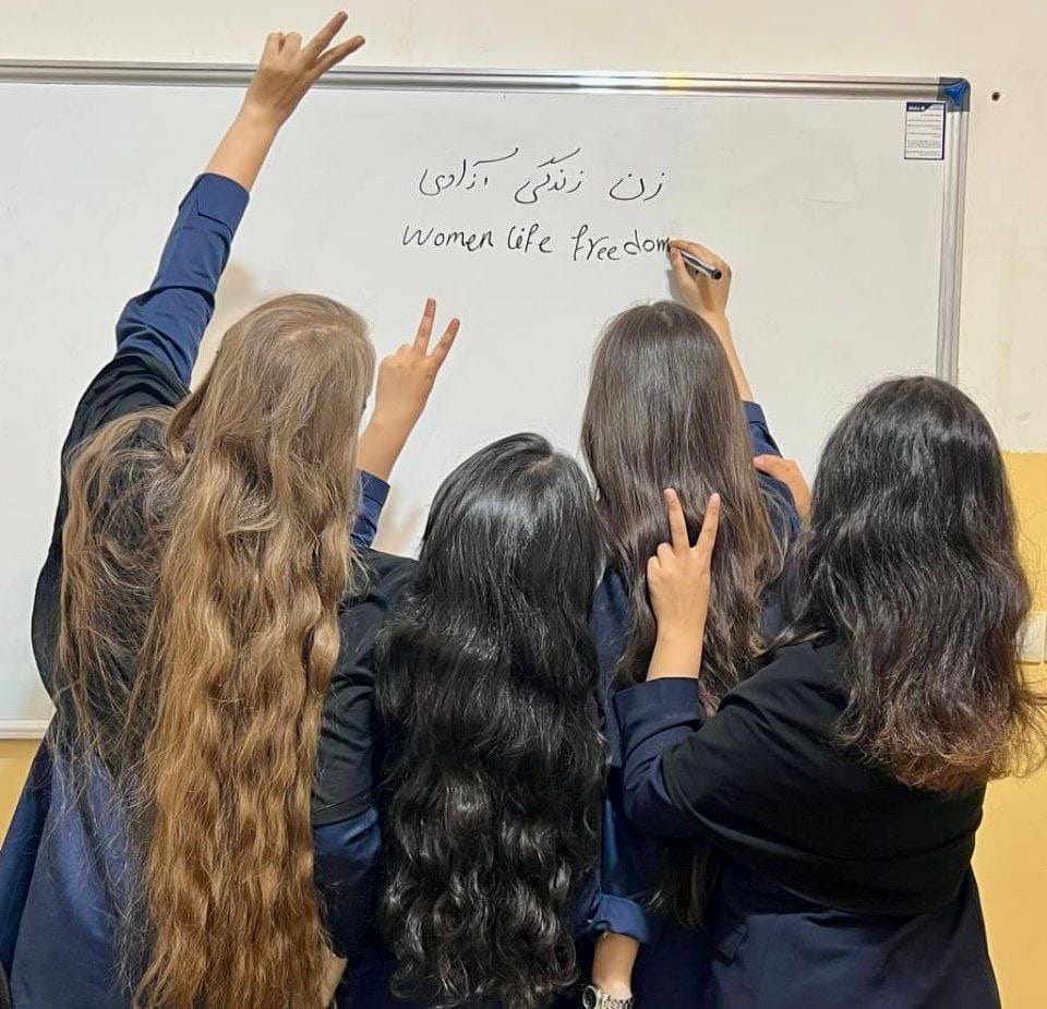 Four Iranian women with uncovered hair stand in front of a whiteboard, raising peace signs and writing "Women, Life, Freedom" in English and Farsi.