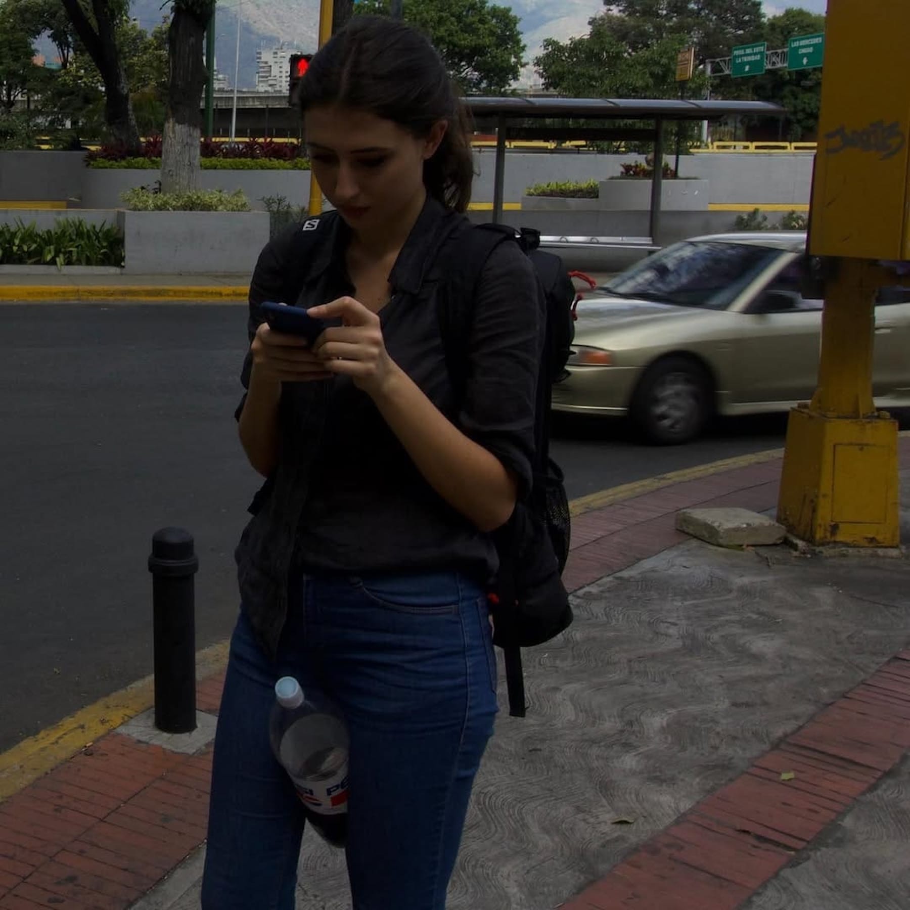 Sala is standing on a sidewalk, focused on her phone. She wears a black button-up shirt, blue jeans, and a backpack. A car, yellow street signs and trees are visible in the background.