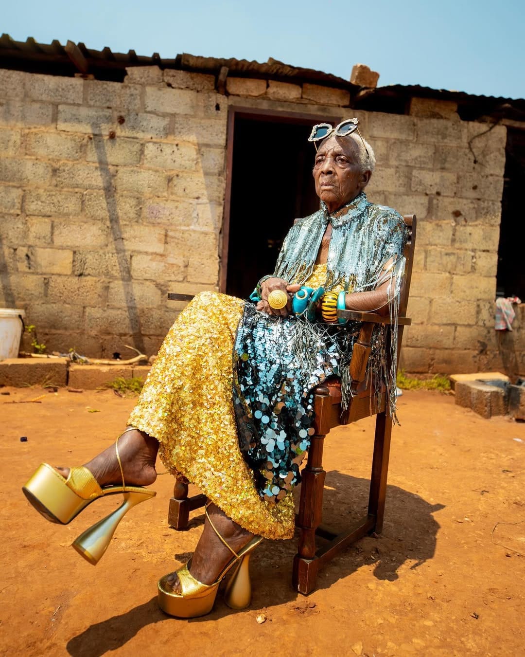 Chola is sitting regally in a wooden chair, wearing a shimmering silver and gold outfit, oversized sunglasses, and high-heeled golden platform shoes. The backdrop features a modest brick house with a rusted roof in Zambia's countryside..