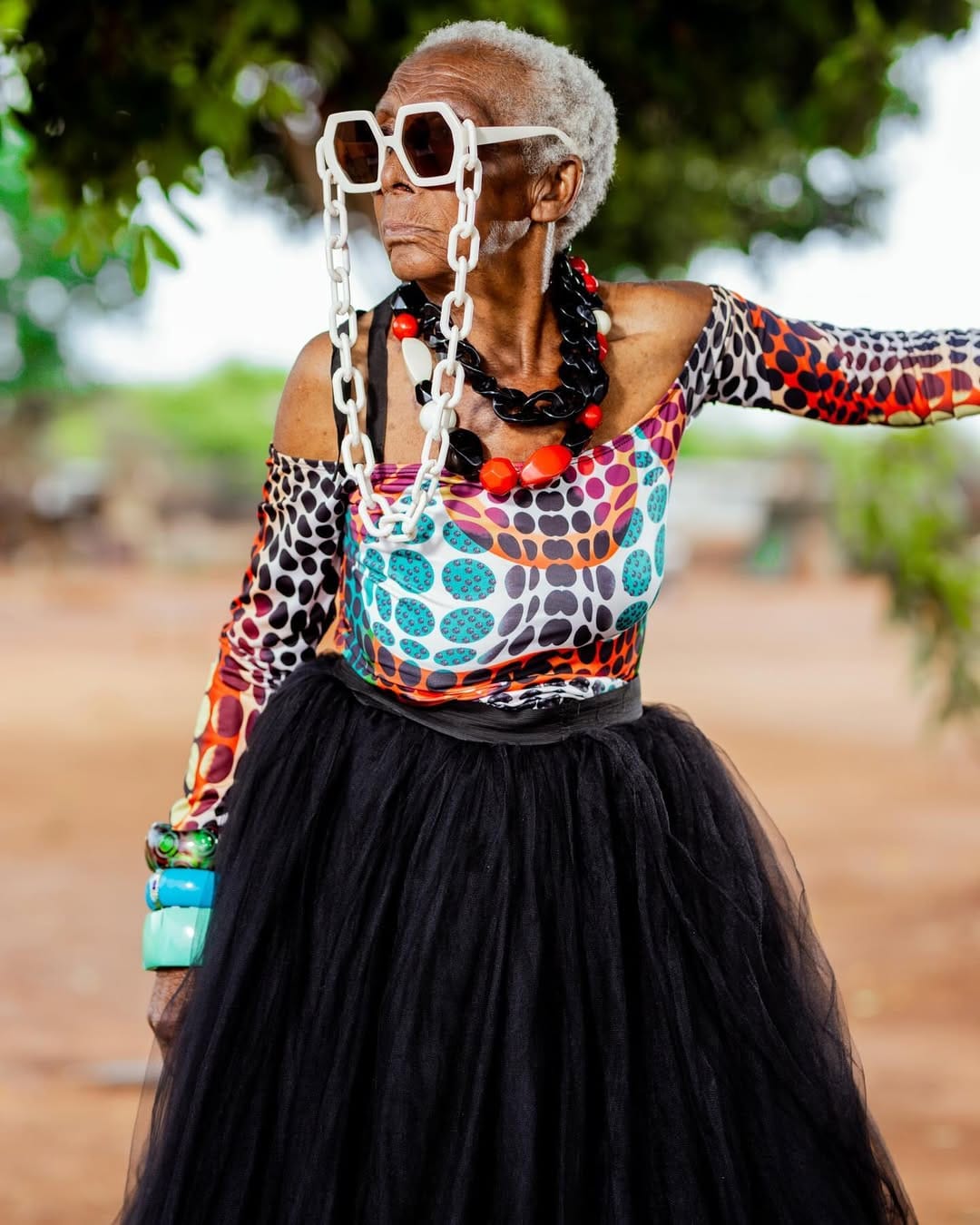 A close shot of Chola posing against a tree, wearing a bold polka-dot top, oversized white chain glasses, and a large black tulle skirt.