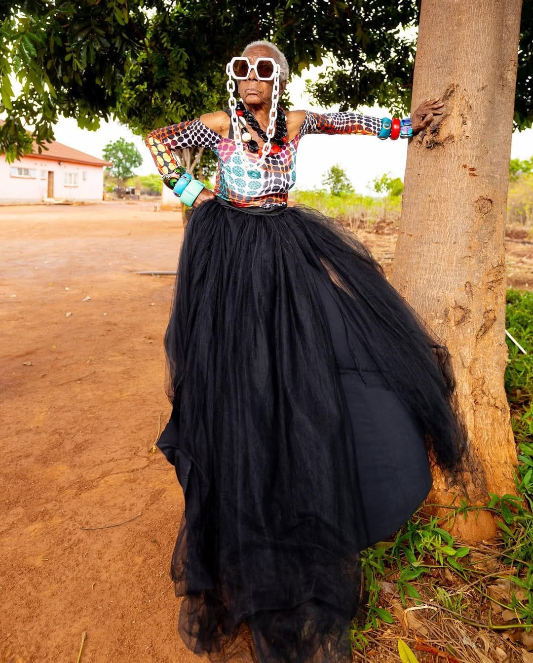 Chola makes a bold pose against a tree in rural setting. She is dressed in a bold polka-dot top, oversized white chain glasses, and a large black tulle skirt.