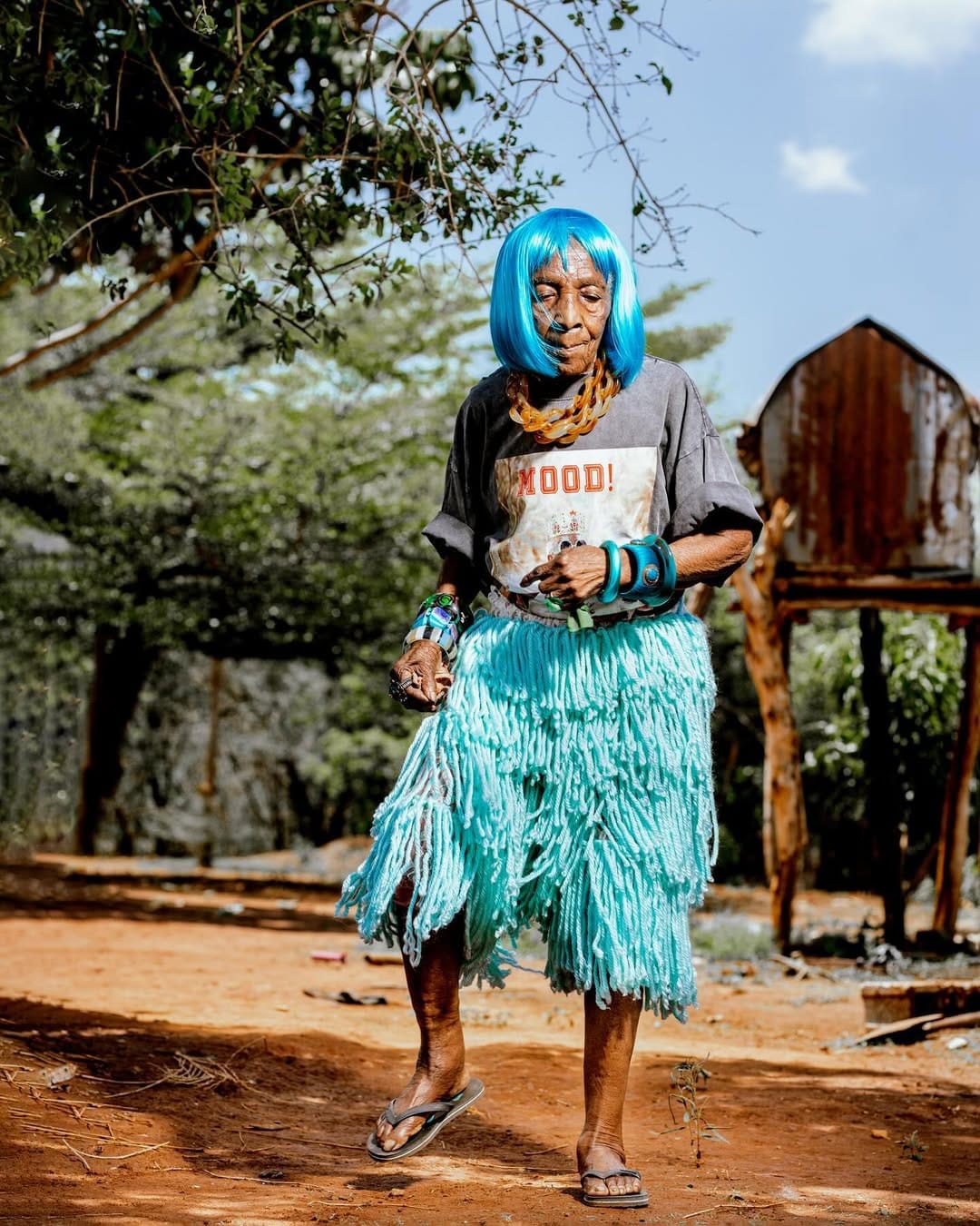 Chola is dancing in small sway moves under some tree shades in a sunlit rural setting of Zambia. She is wearing a bold, bright blue wig, heavy-sized amber chain necklaces, a modern T-shirt, a light blue grass skirt, and a pair of slippers.