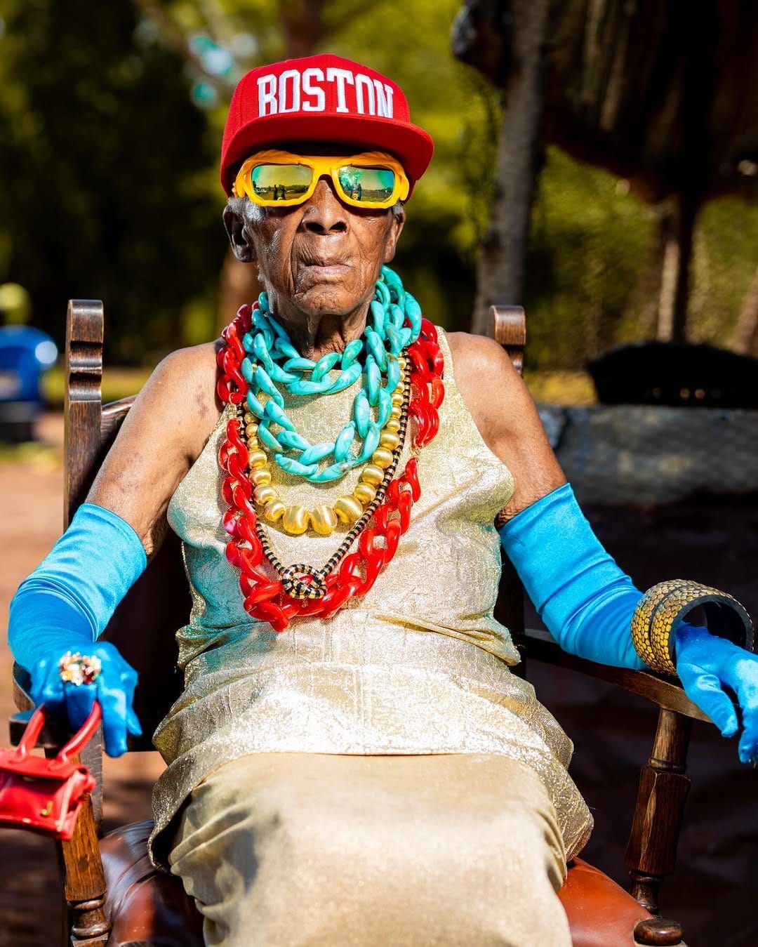 Chola sits confidently in a chair with calm expression, a simple gold dress, oversized catchy blue gloves, chunky colorful jewelry, a red "Boston" cap, and reflective yellow sunglasses.