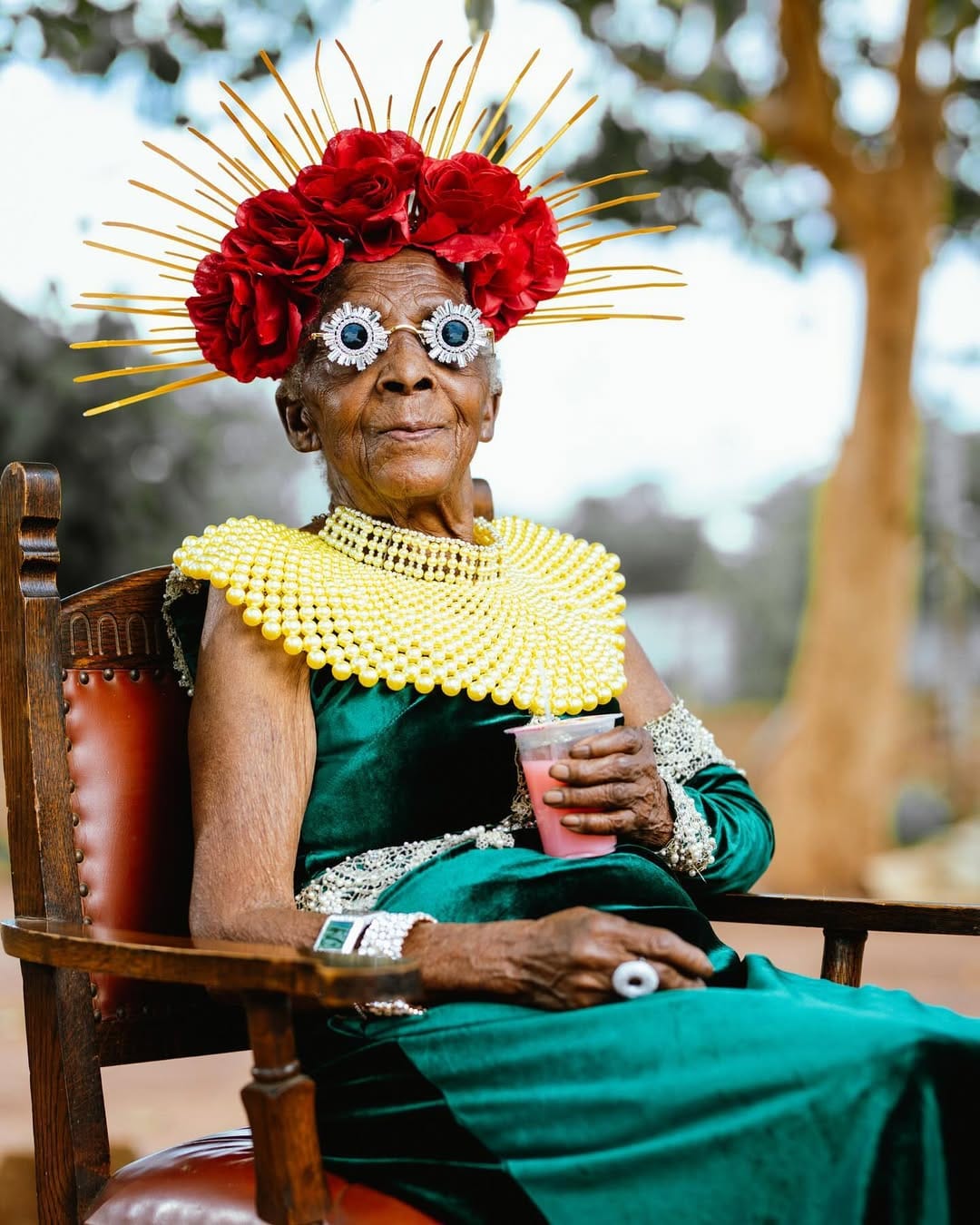 Chola is sitting in a wooden chair and wearing a striking emerald green gown. She holds a pink beverage and has an elaborate, huge red floral headpiece with golden spikes on her head, eccentric, silver eyewear and a flapped, yellow beaded neckwear.