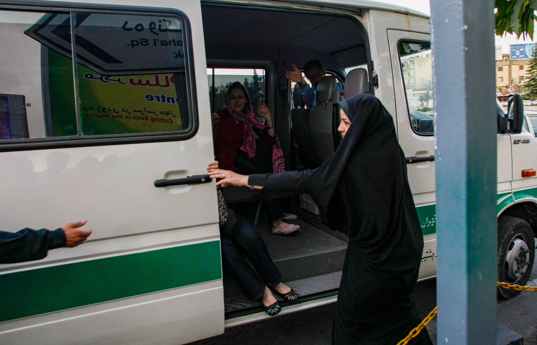 An Iranian woman in a black chador closes the door of a police van as other women sit inside, appearing to be monitored for compliance with dress code regulations. 