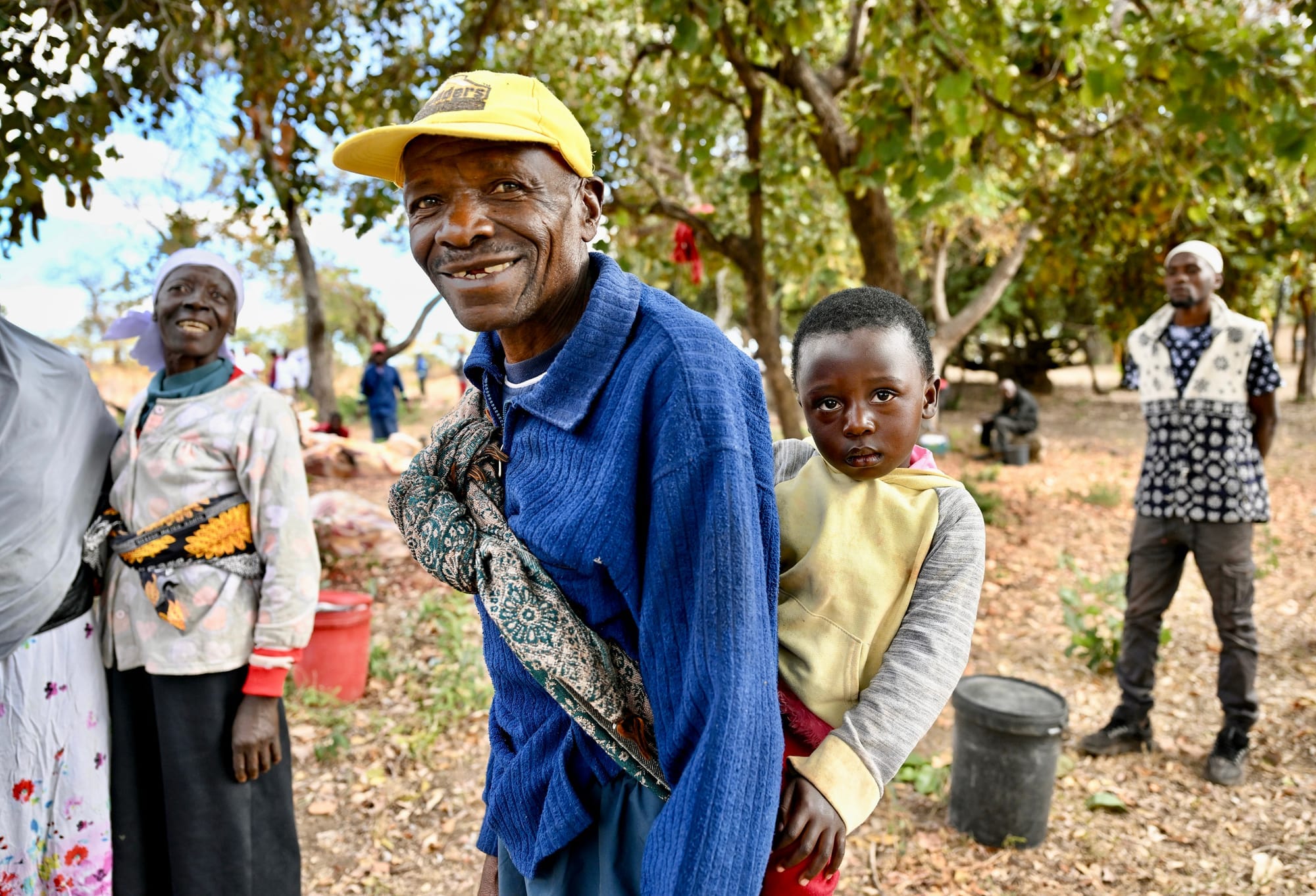 A smiling grandfather in a yellow cap carries his grandchild on his back in Mutoko, Zimbabwe, with other family members and trees in the background.