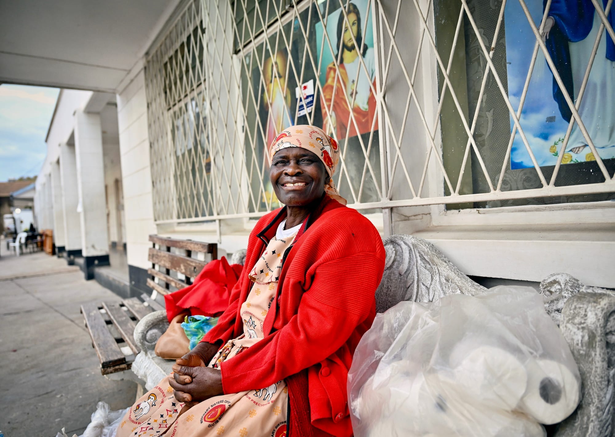 A smiling woman in vibrant red coat sits on a bench in Harare, Zimbabwe, with religious artwork visible and a bag of tissue rolls behind her and near a marketplace.