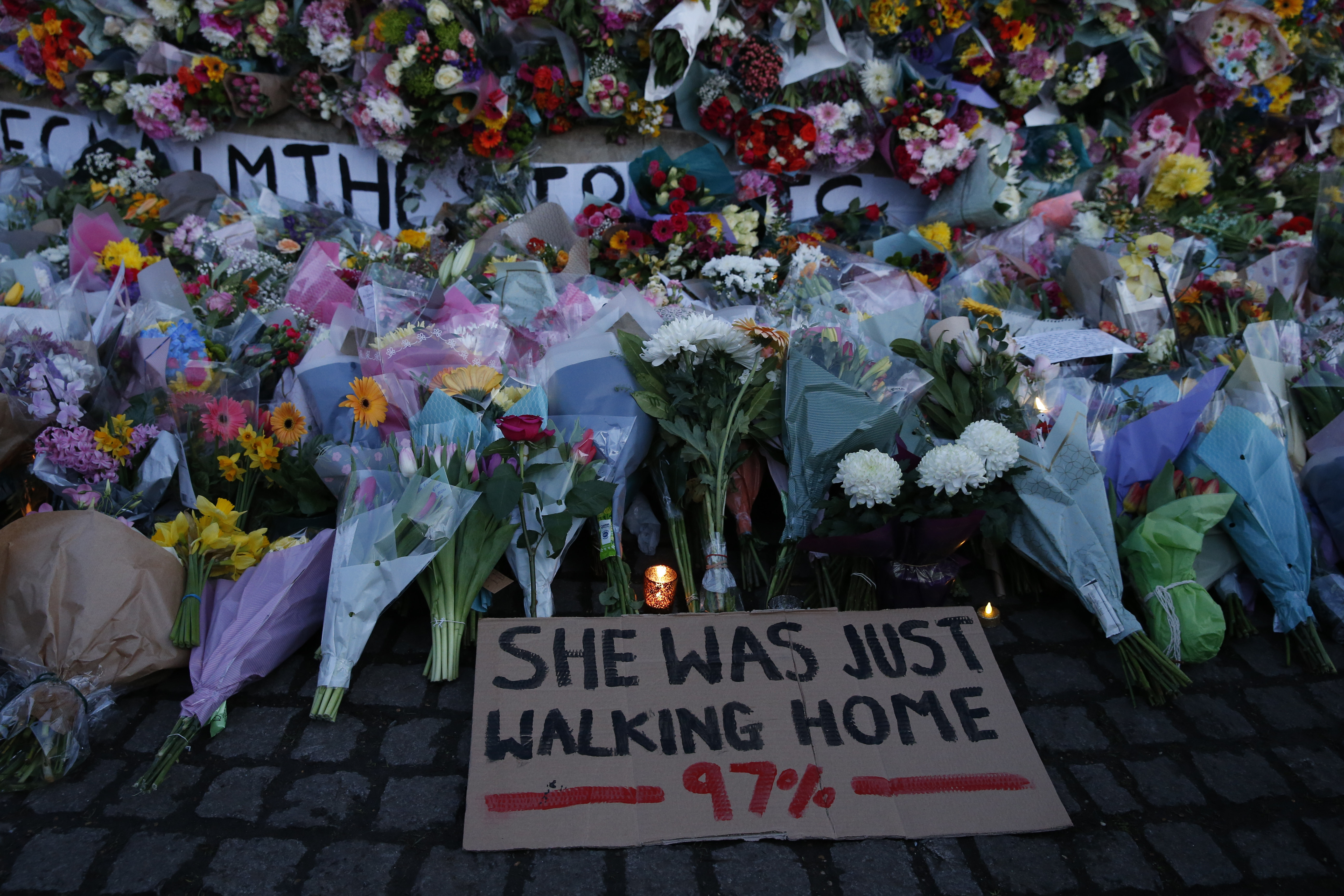 A sign saying "SHE WAS JUST WALKING HOME 97%" with flowers and candles around photo.