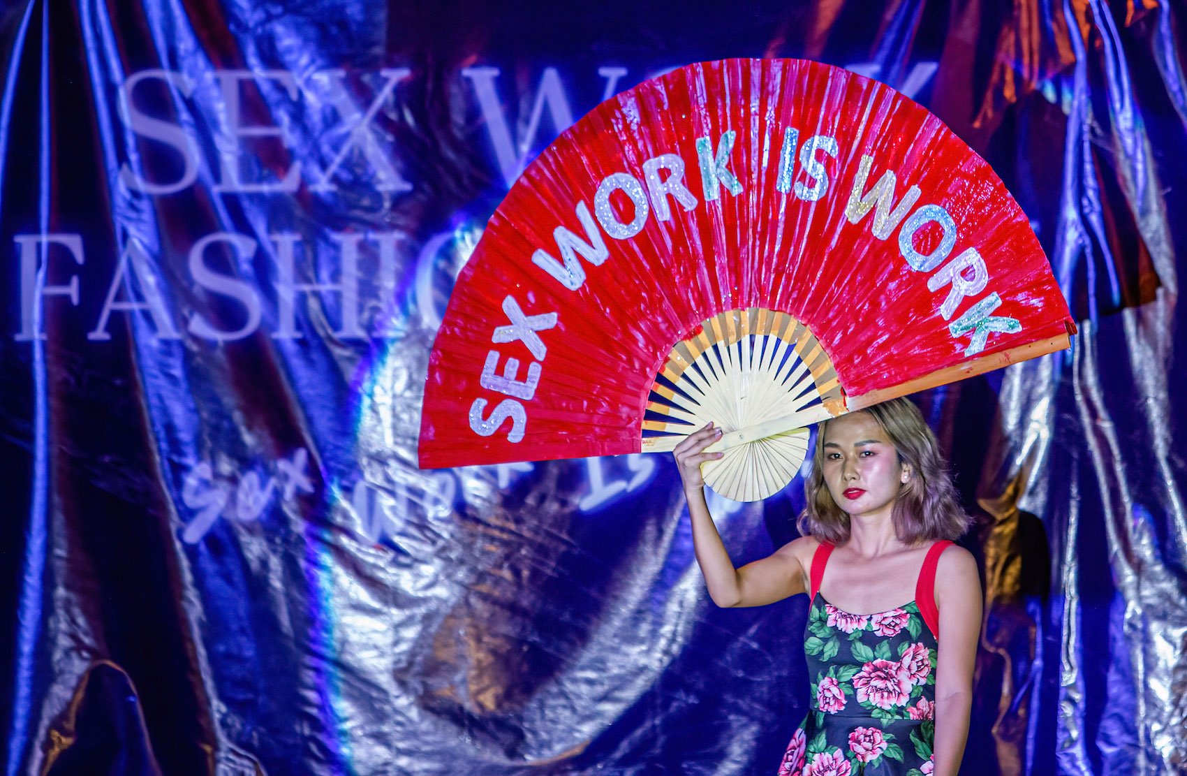 An activist holds a fan shape placard that says "Sex Work Is Work" during a Sex Work Fashion Week on International Labor Day at the Tha Phae Gate