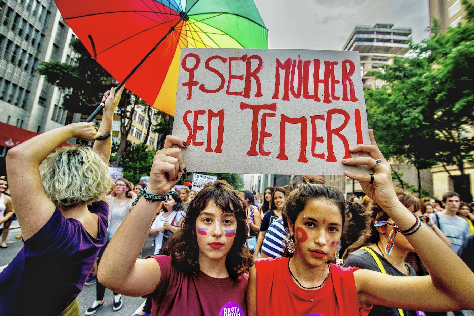 A protest sign is held by two women during a march in Brazil. A rainbow umbrella is held up behind them.