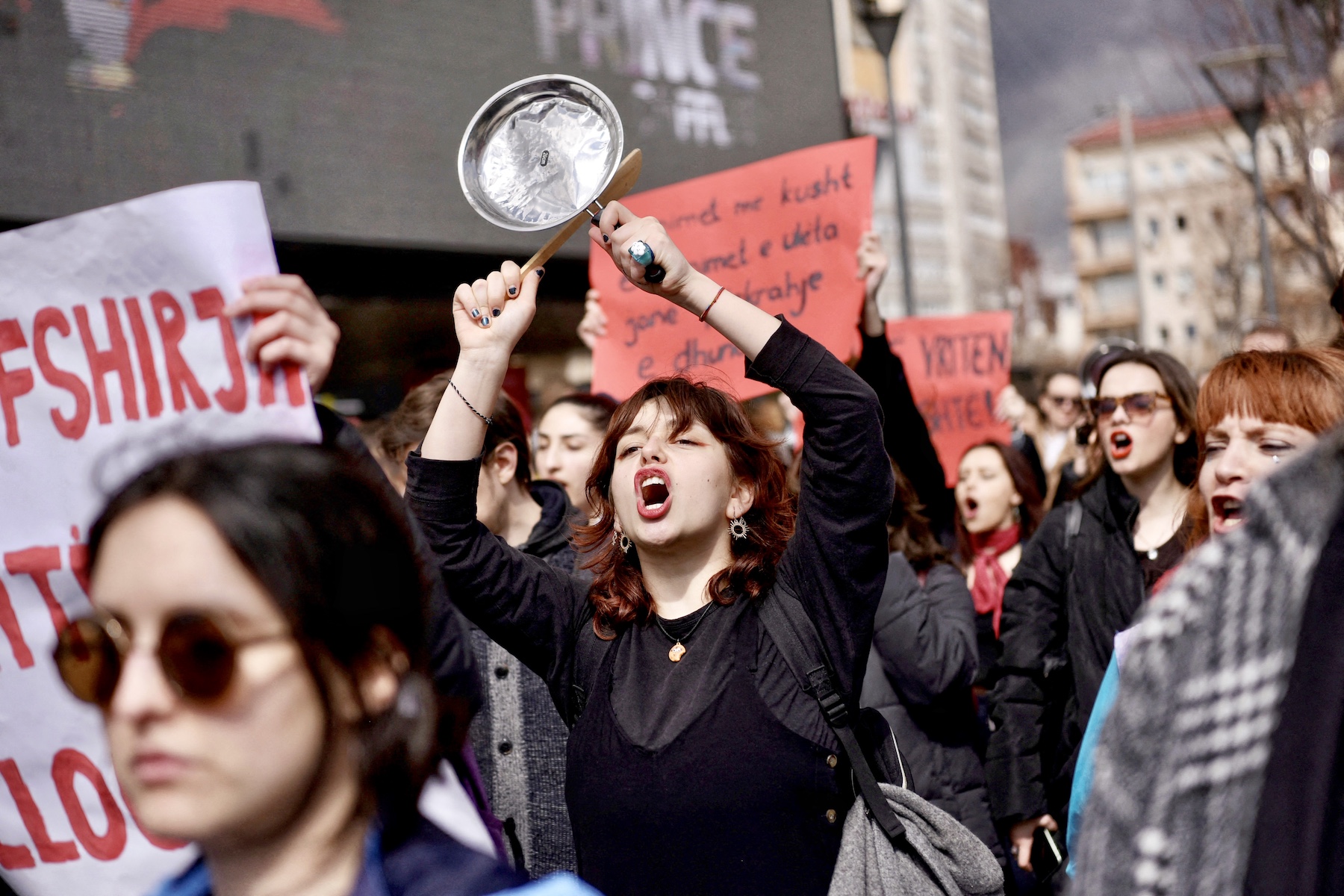 Woman makes noise with a pan and wooden spoon at the International Women's Day march in Kosovo. She is surrounded by women, who also appear to be shouting and holding red banners.