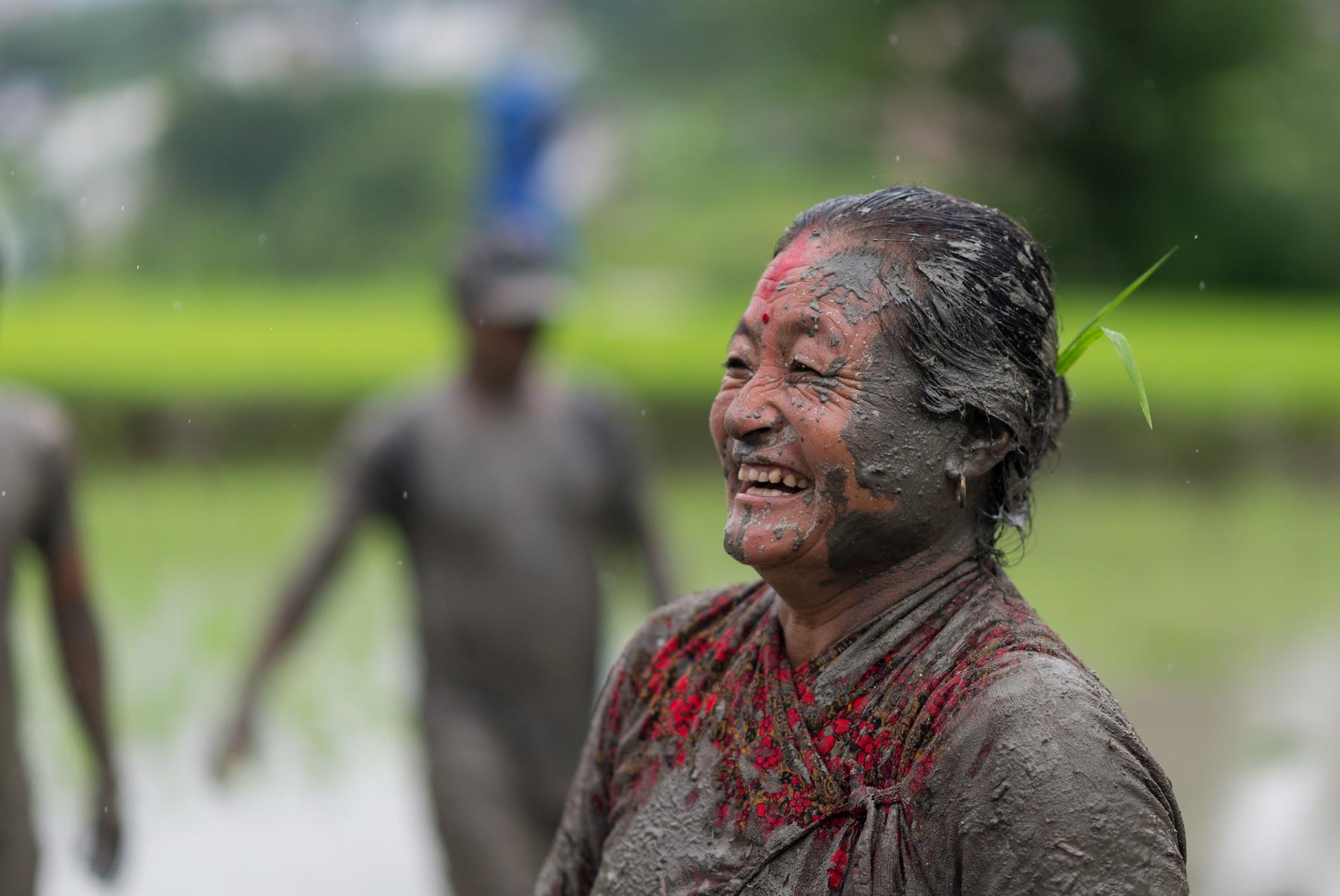 A woman, who appears to be older, smiles while her face and clothes are covered in mud.