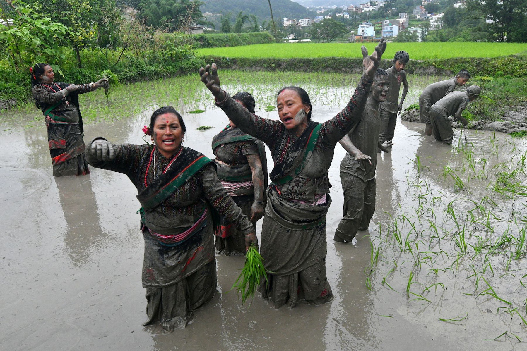 Two women dance in a rice paddy field. They are covered in mud. People in a similar state are seen in the back.