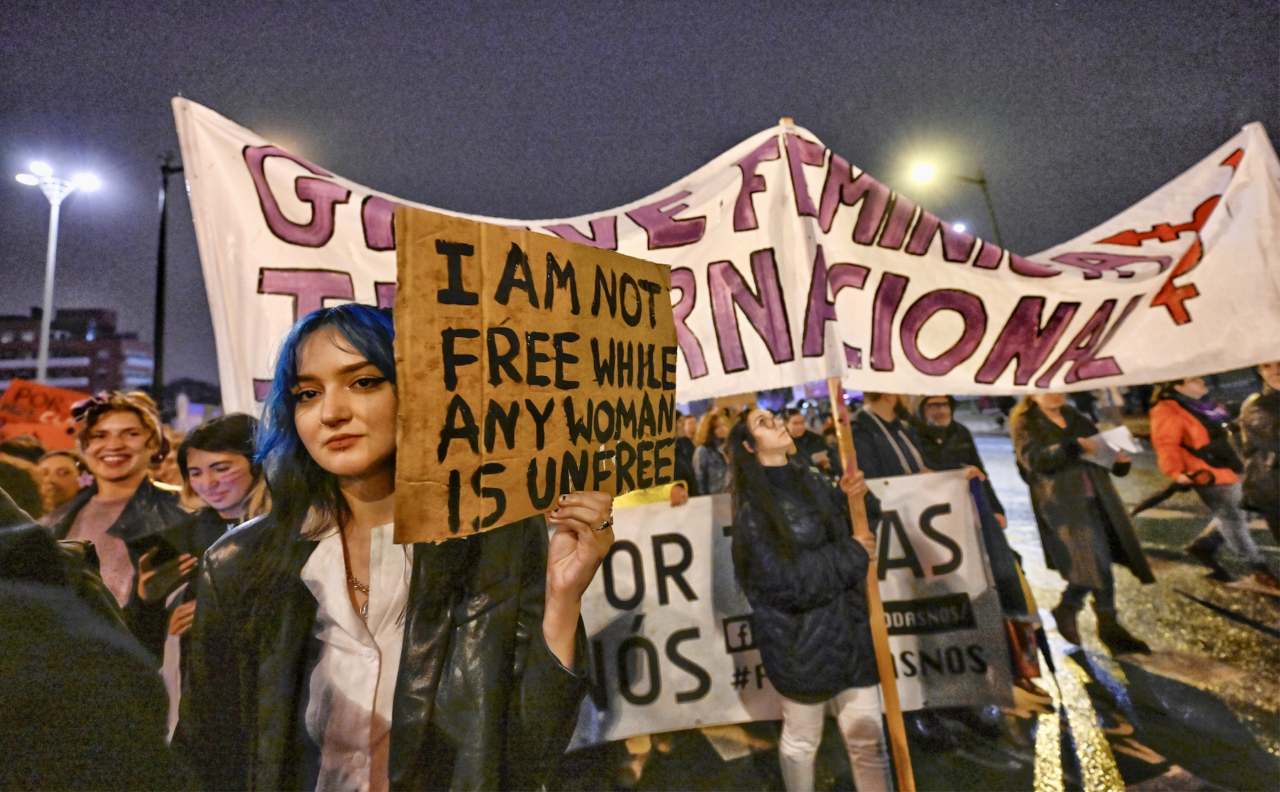 A woman with blue hair holds up a cardboard sign that says, "I am not free while any women is unfree". 