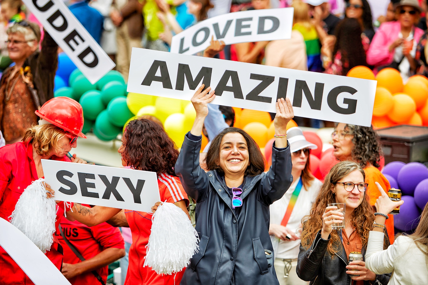 A woman holds a sign that says, "Amazing". Around her people hold other signs that read, "Sexy" and "Loved".